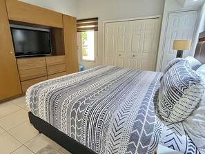 A bedroom with a bed, a television mounted on a wooden cabinet, a white door, and a window with blinds. - Playa Royale Beachfront Family Condo °BBQ, Terrace & Garden Views° (Nuevo Nayarit)