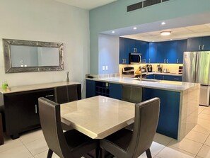 A modern kitchen with a central island, dark cabinets, and a dining area with a table and chairs. - Playa Royale Beachfront Family Condo °BBQ, Terrace & Garden Views° (Nuevo Nayarit)