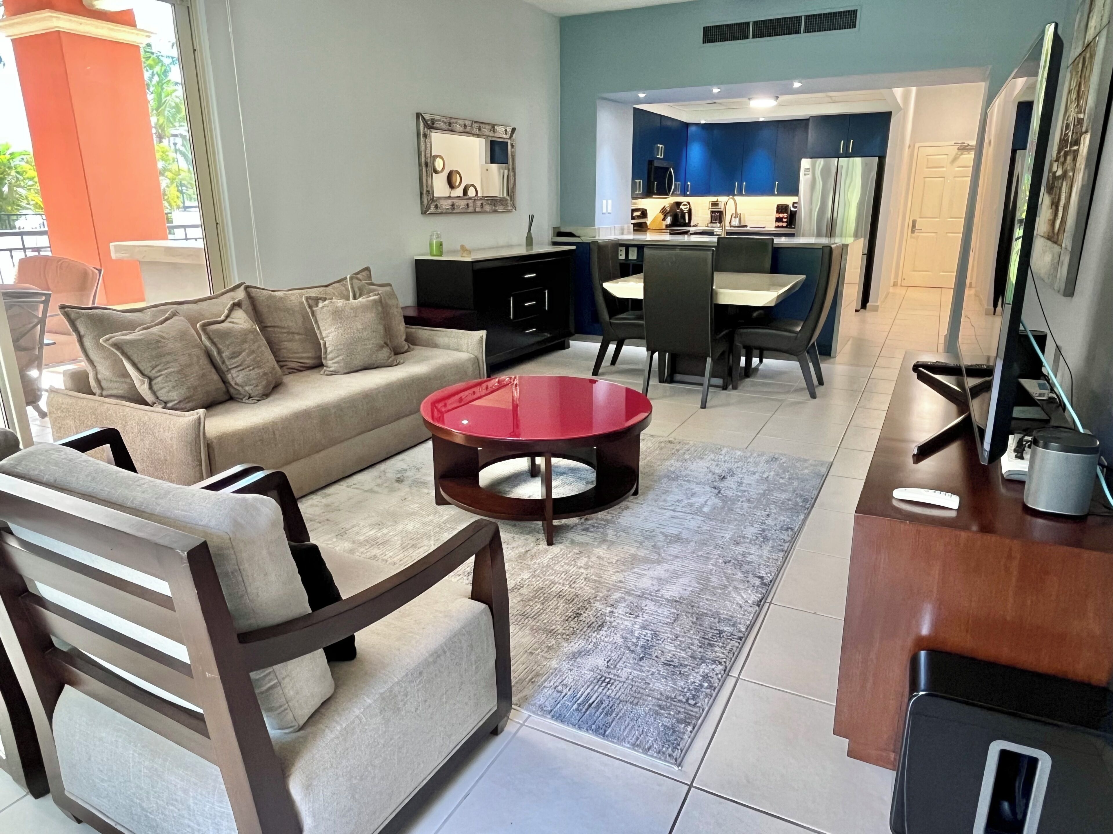 A modern living room with a sofa, a red coffee table, and a dining area with chairs and a kitchen in the background.