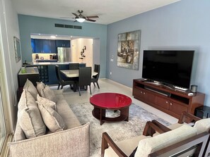 A living room with a sofa, a red coffee table, a TV, and a dining area with chairs and a kitchen in the background. - Playa Royale Beachfront Family Condo °BBQ, Terrace & Garden Views° (Nuevo Nayarit)