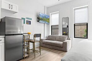 Et moderne køkken med et køleskab, et spiseområde med stol og bord, en sofa og et stort vindue med udsigt til en murstensbygning.