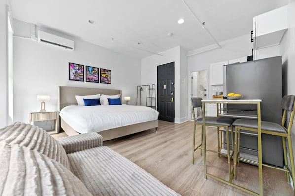 A modern hotel room with a bed, bedside tables, a desk with a chair, a refrigerator, and artwork on the wall.