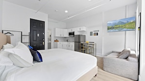 A modern hotel room with a bed, a seating area, a kitchenette, and a TV mounted on the wall.