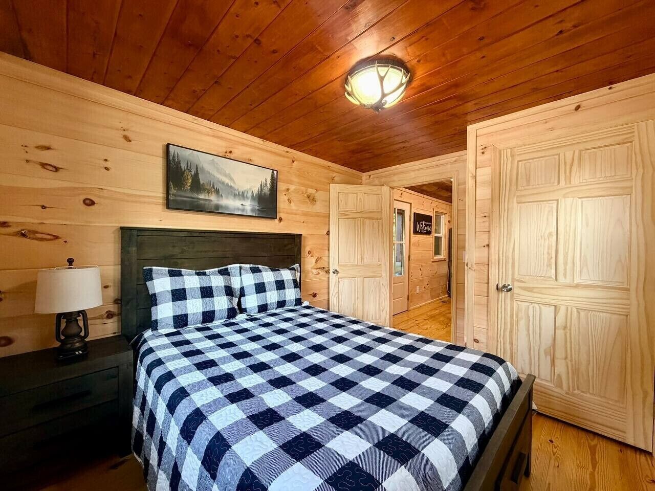 A wooden cabin bedroom with a checkered bedspread, a nightstand with a lamp, and a painting on the wall.