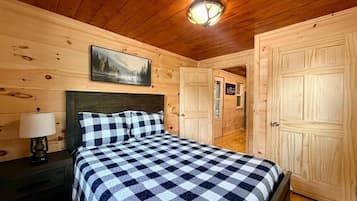 A wooden cabin bedroom with a checkered bedspread, a nightstand with a lamp, and a painting on the wall.
