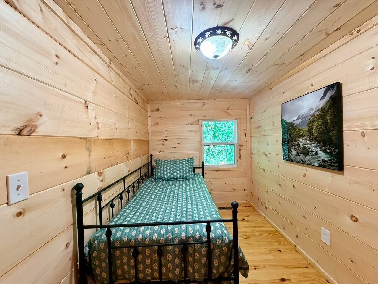 Una habitación pequeña con cama individual, interior de madera, ventana con vistas a la vegetación y un cuadro en la pared.