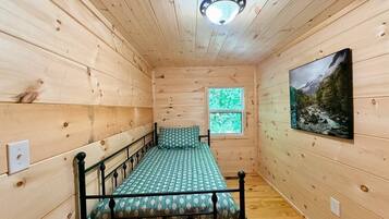 Una habitación pequeña con cama individual, interior de madera, ventana con vistas a la vegetación y un cuadro en la pared.