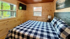Un dormitorio de cabaña de madera con colcha a cuadros, dos ventanas, un televisor montado y un cuadro en la pared.