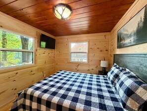 Quarto de cabana de madeira com colcha xadrez, duas janelas, televisão na parede e um quadro.