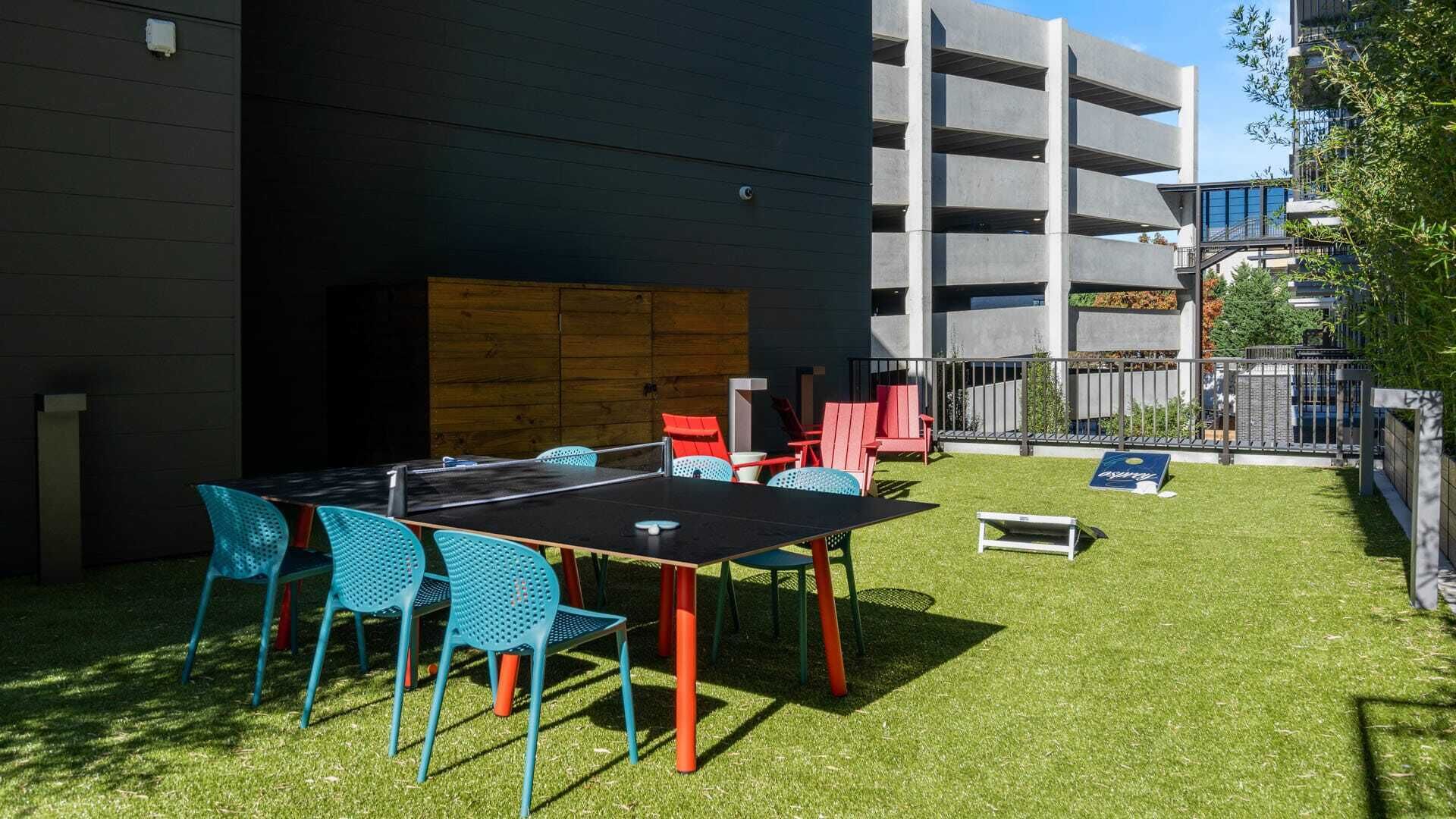 Un espace extérieur aménagé avec une table de ping-pong, des chaises et une petite table, entouré d’une pelouse verdoyante et d’un bâtiment à la façade sombre.