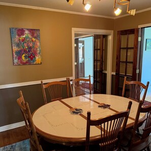 Uma sala de jantar com uma mesa redonda, cadeiras de madeira, um quadro colorido e uma porta que dá para o exterior.