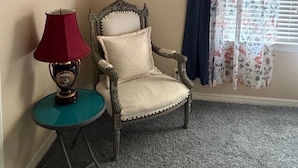 Una habitació amb una cadira, una taula amb una làmpada, un quadre amb marc a la paret i una finestra amb cortines.