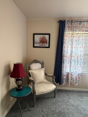 Ein Zimmer mit einem Stuhl, einem Tisch mit Lampe, einem gerahmten Bild an der Wand und einem Fenster mit Vorhängen.