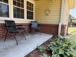 Eine Veranda mit zwei Stühlen, einem kleinen Tisch, einer Ziegelmauer und einer Pflanze.
