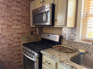 Una cocina con pared de ladrillo, electrodomésticos de acero inoxidable, encimeras de granito y una ventana con persianas.