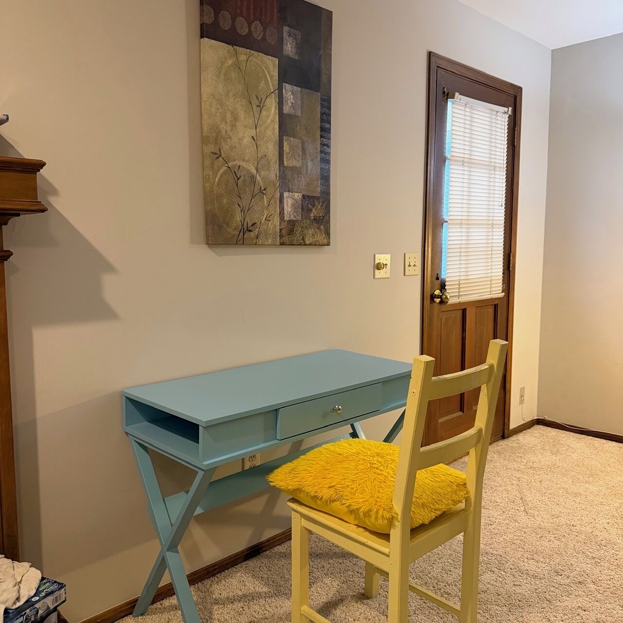 A room with a blue desk, a wooden chair with a yellow cushion, a door with blinds, and a wall-mounted artwork.