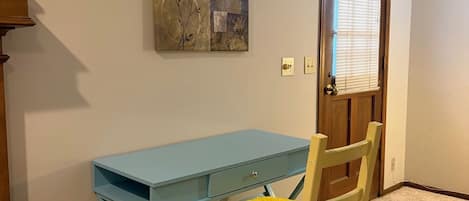 A room with a blue desk, a wooden chair with a yellow cushion, a door with blinds, and a wall-mounted artwork.