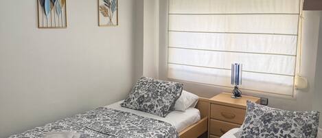 A bedroom with two beds, a ceiling fan, a window with blinds, and artwork on the wall.