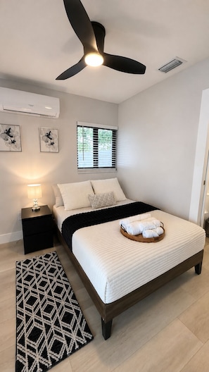 A bedroom with a bed, a bedside table, a floor lamp, a ceiling fan, a window with blinds, and a wall-mounted air conditioner. - The Lincoln Luxe Studio• Private Retreat • Midtown • Downtown • Stadium  (Tampa)