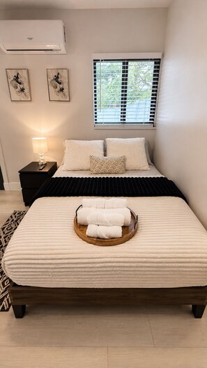 A bedroom with a bed, a bedside table with a lamp, a window with blinds, and an air conditioner on the wall. - The Lincoln Luxe Studio• Private Retreat • Midtown • Downtown • Stadium  (Tampa)