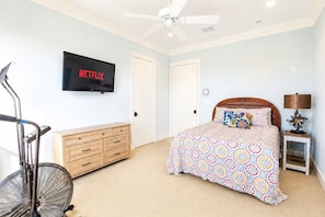 A bedroom with a bed, a TV mounted on the wall, a wooden dresser, a treadmill, and a standing fan. - Beachfront + Canal: 4BR/4BA w/ Ocean Views & Fireplace (Port Bolivar)