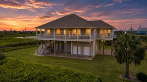 A two-story house with a balcony, a covered porch, and a staircase leading to the upper level. - Beachfront + Canal: 4BR/4BA w/ Ocean Views & Fireplace (Port Bolivar)