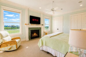 A bedroom with a fireplace, a bed, a television mounted above it, and a view of the outdoors through a window. - Beachfront + Canal: 4BR/4BA w/ Ocean Views & Fireplace (Port Bolivar)