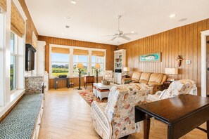 A spacious living room with a large window, a ceiling fan, and a view of the outdoors. - Beachfront + Canal: 4BR/4BA w/ Ocean Views & Fireplace (Port Bolivar)