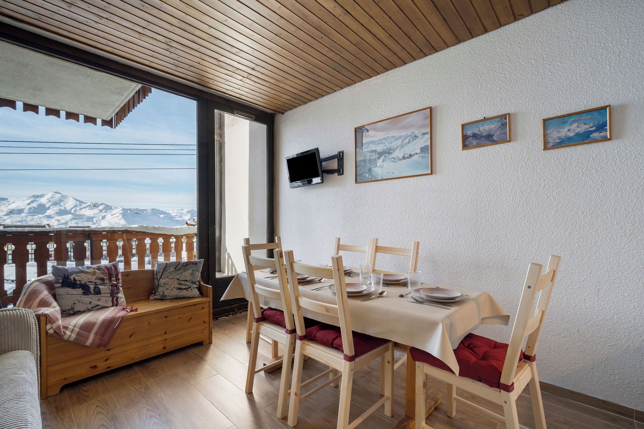 Un'area pranzo con tavolo imbandito per quattro persone, sedie in legno, una panca con cuscini e vista sulle montagne innevate attraverso una finestra.
