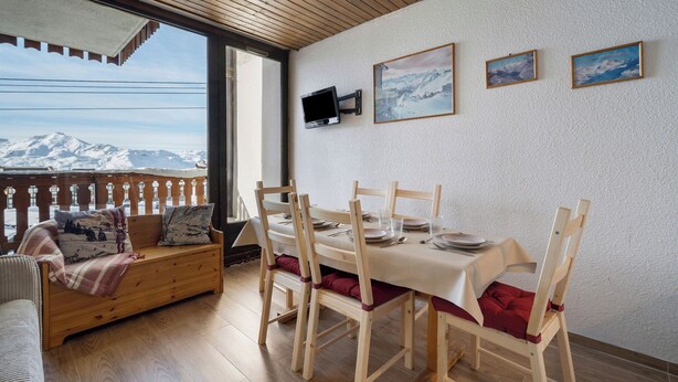 A dining area with a table set for four, wooden chairs, a bench with cushions, and a view of snow-capped mountains through a window. - Résidence les Eterlous (Les Belleville)