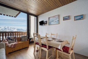 A dining area with a table set for four, wooden chairs, a bench with cushions, and a view of snow-capped mountains through a window. - Résidence les Eterlous (Les Belleville)