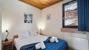 A bedroom with a bed, a nightstand, a lamp, a window with a view of a snowy landscape, and a framed picture on the wall. - Résidence les Eterlous (Les Belleville)