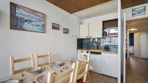 A dining area with a table set for four, wooden chairs, and a kitchen with white cabinets and a built-in oven. - Résidence les Eterlous (Les Belleville)