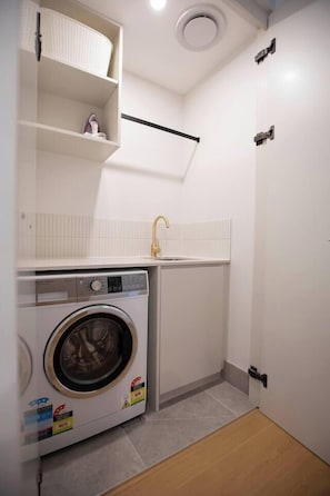 A compact laundry room with a washing machine, a sink, and a shelf with shoes. - The Aristocrat | Mid Century Luxe Dual Suite + Gym (Dickson)