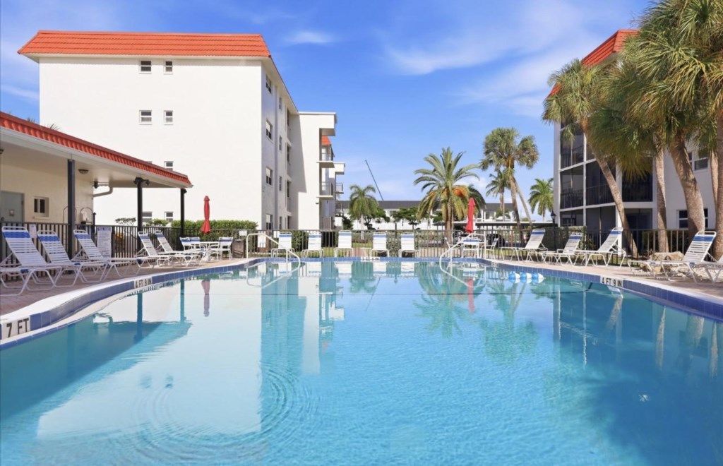 Una piscina con sillones de descanso, un edificio blanco con techo rojo y palmeras.