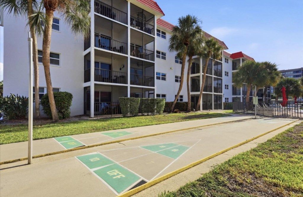 A multi-story residential building with balconies, a parking area with marked spaces, and palm trees.