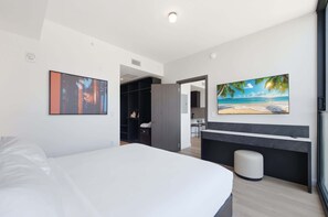 Ein modernes Hotelzimmer mit einem Bett, einem Schreibtisch, einem Stuhl, einem kleinen Hocker, einem Kleiderschrank und einem Gemälde mit einer Strandansicht.