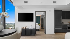 Ein modernes Wohnzimmer mit Flachbildfernseher, Essbereich mit Tisch und Stühlen sowie eine Küche mit dunklen Schränken und einer Kaffeemaschine.