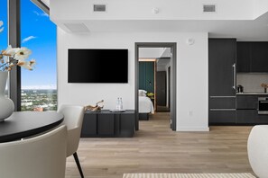 Ein modernes Wohnzimmer mit Flachbildfernseher, Essbereich mit Tisch und Stühlen sowie eine Küche mit dunklen Schränken und einer Kaffeemaschine.
