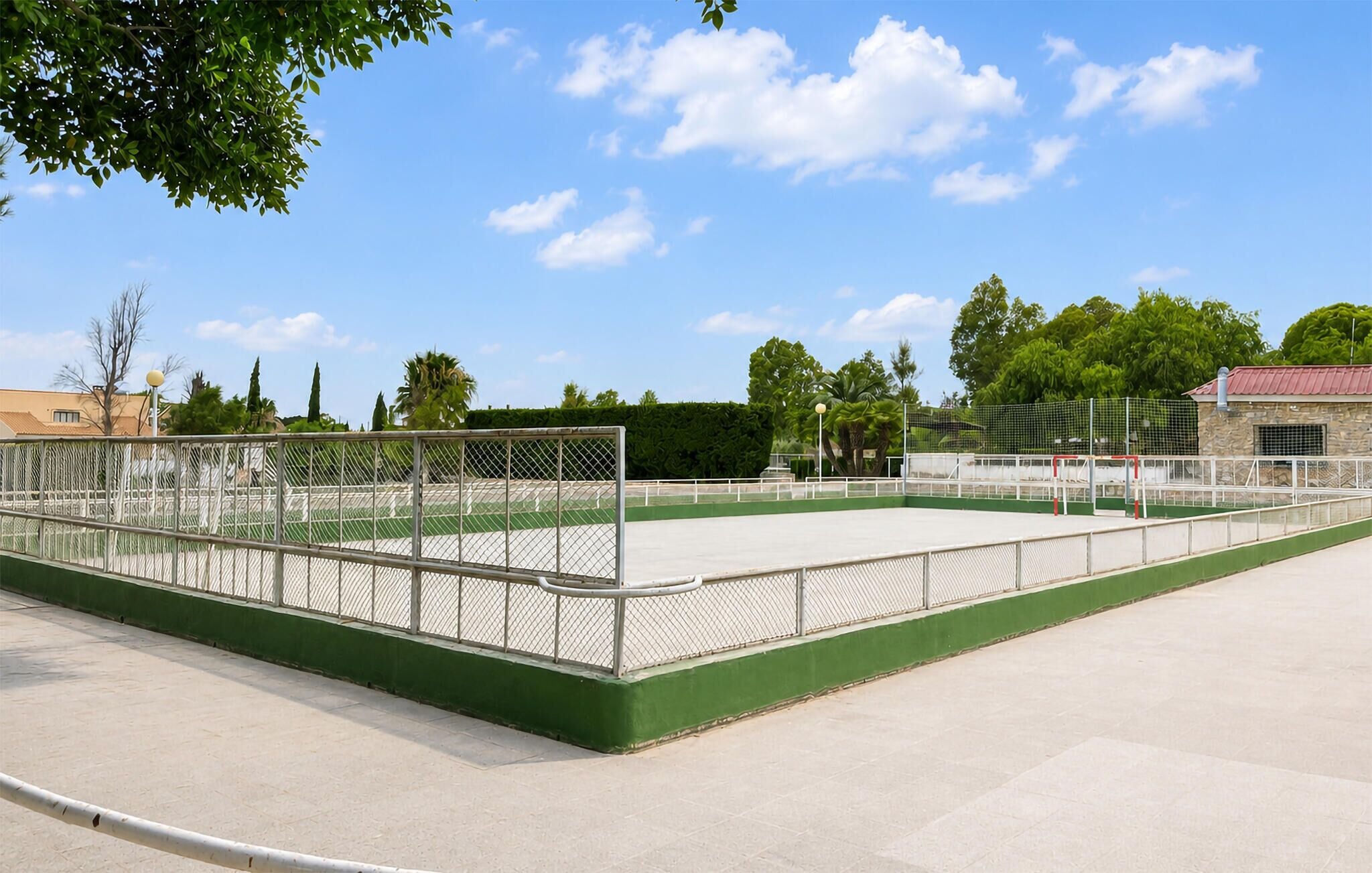 An outdoor tennis court with a chain-link fence, green flooring, and a clear sky.