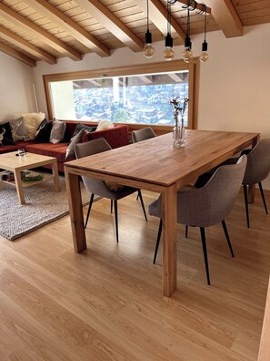 A dining area with a wooden table and chairs, a sofa with cushions, a rug, and a view through a large window. - Luxury Moments Begin, Wallis 1077 (Wallis)