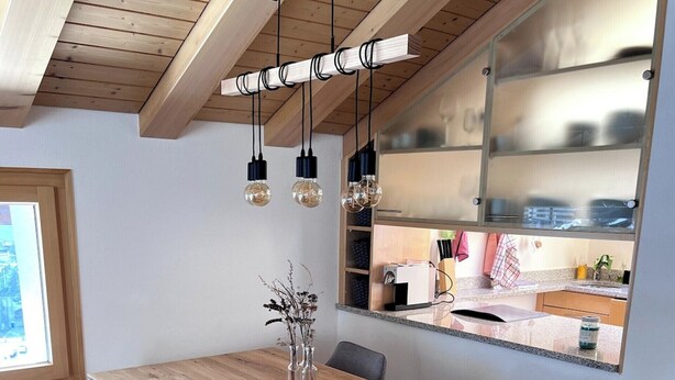 A dining area with a wooden table, grey upholstered chairs, hanging pendant lights, and a kitchen area with a stainless steel backsplash. - Luxury Moments Begin, Wallis 1077 (Wallis)