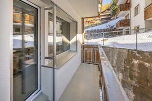 Ein schmaler Korridor mit einer Glastür zu einem Raum, einem Holzzaun und einem schneebedeckten Hang mit Gebäuden im Hintergrund.