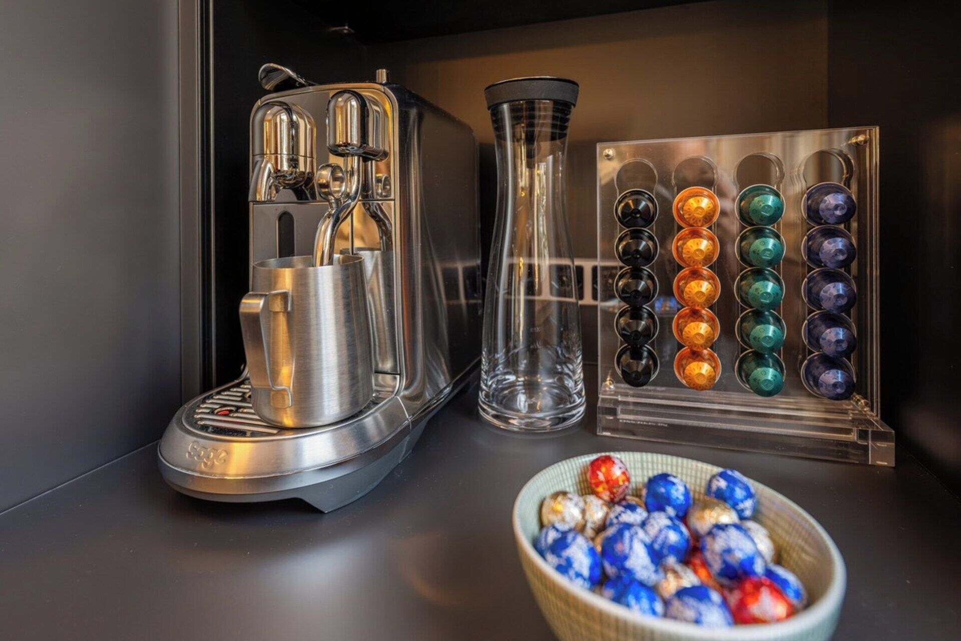Uma máquina de café, uma jarra de vidro e um conjunto de cápsulas coloridas em uma bancada.