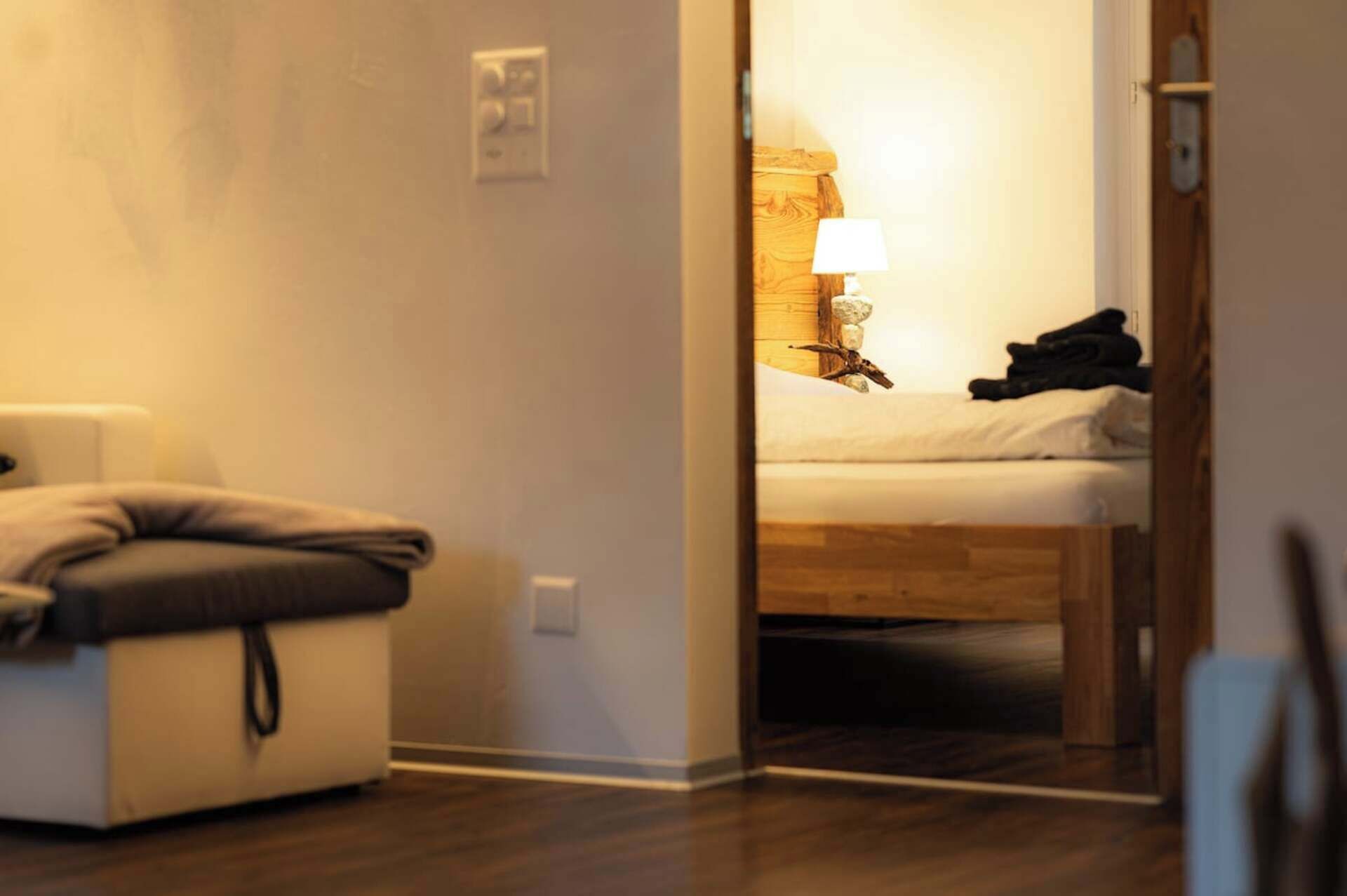 A hotel room with a bed, a bedside table with a lamp, and a small seating area with a cushion.