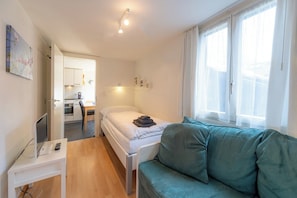 Ein Schlafzimmer mit einem Bett, einem Schreibtisch mit Computer, einem Fenster mit Vorhängen und Blick auf eine Küche.