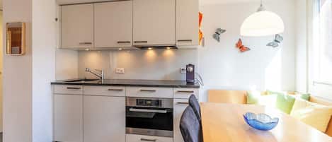 Una cocina moderna con armarios blancos, horno empotrado y comedor con mesa y sillas de madera.