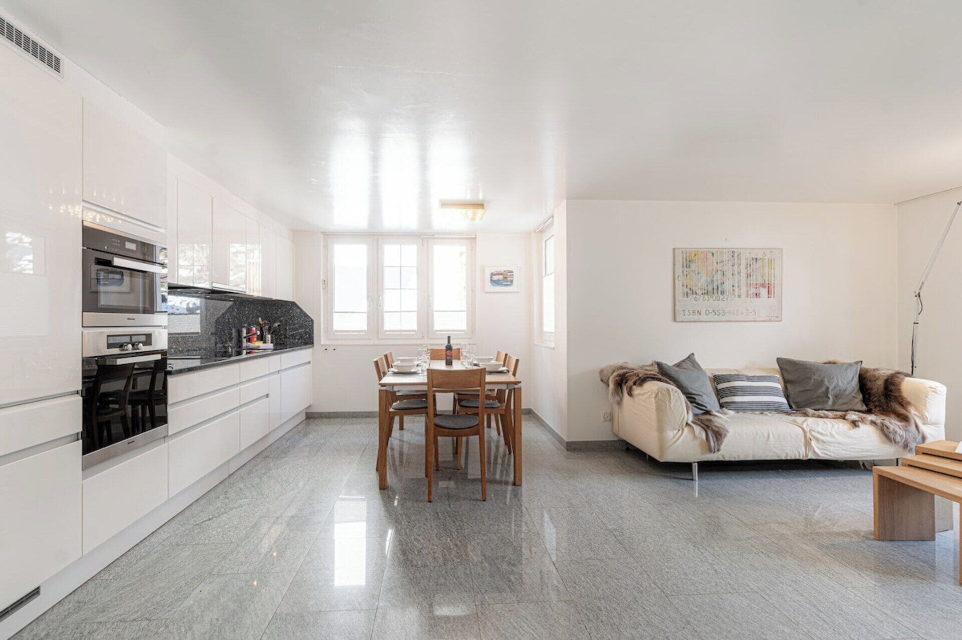 A modern kitchen with white cabinets, a dining table with chairs, and a sofa in a living area with a window.