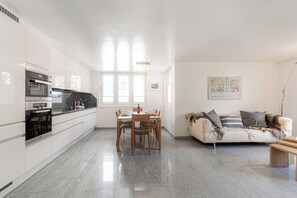 Una cocina moderna con armarios blancos, una mesa de comedor con sillas y un sofá en una sala de estar con ventanal.