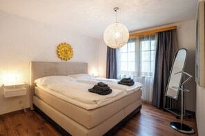 A hotel room with a large bed, a bedside lamp, a sun-shaped wall decoration, a hanging light fixture, and a mirror. - Stay in Alpine Elegance, Wallis 1076 (Wallis)